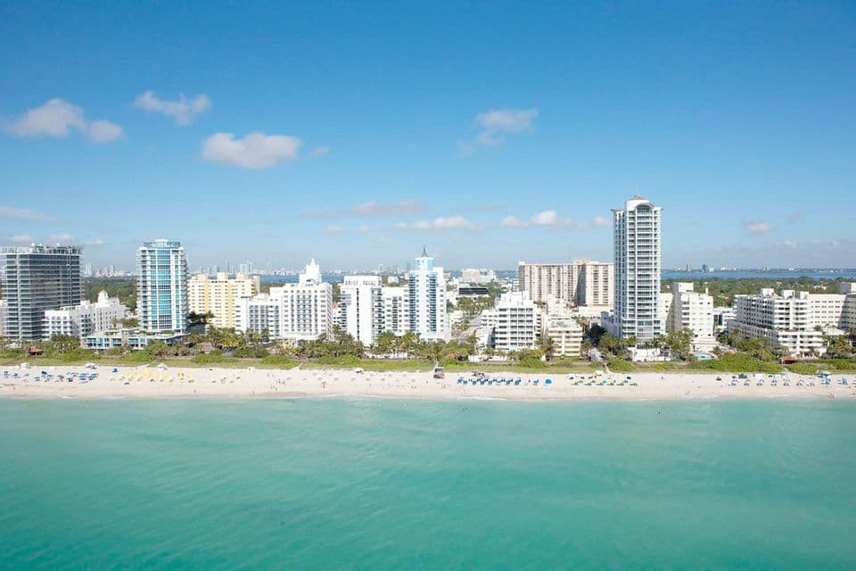 A cityscape of tall buildings lines a white sand beach next to turquoise ocean water under a clear blue sky.