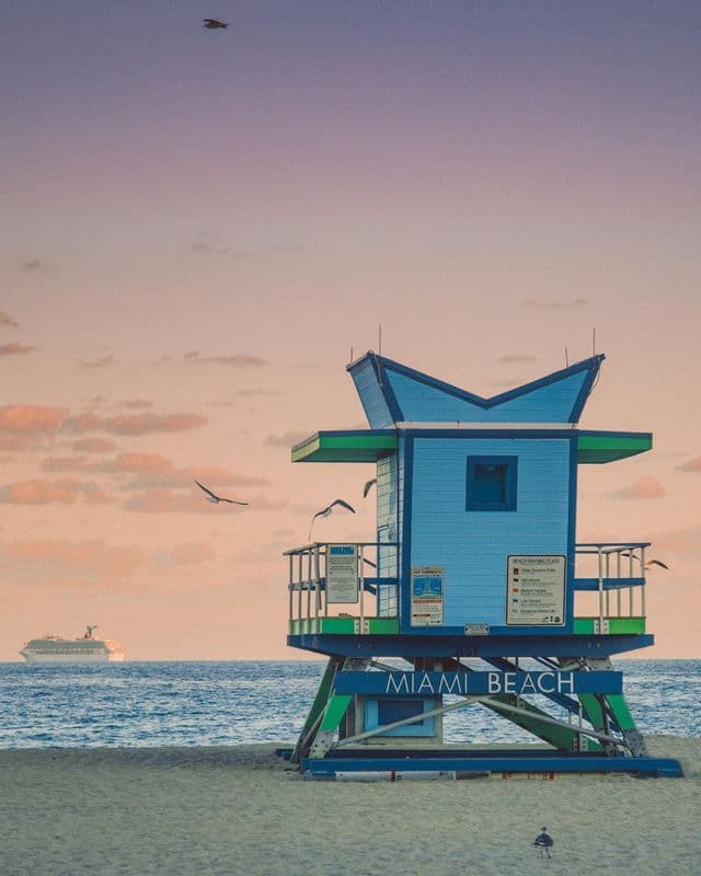 A blue and green Miami Beach lifeguard tower stands on the sand with a cruise ship on the ocean and seagulls in a pastel sky.