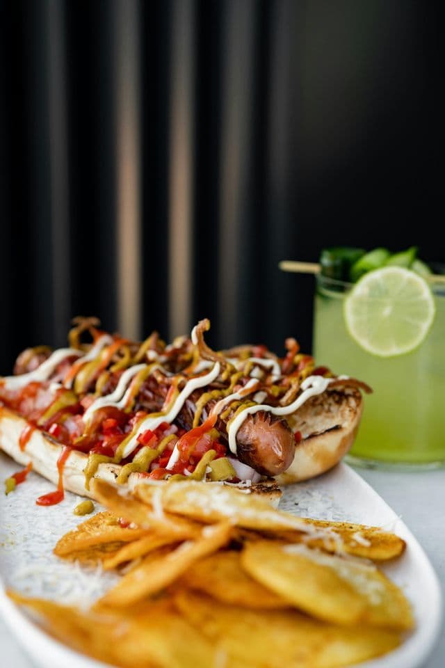 A loaded hot dog with various sauces on a plate with chips and a green cocktail garnished with lime.