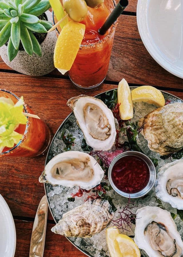 A top-down view of a platter of fresh oysters on ice with lemon and sauce, next to two Bloody Mary cocktails on a wooden table.