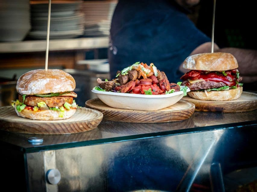 Two burgers and a bowl of meat and vegetables are displayed on wooden plates on a food stall counter.