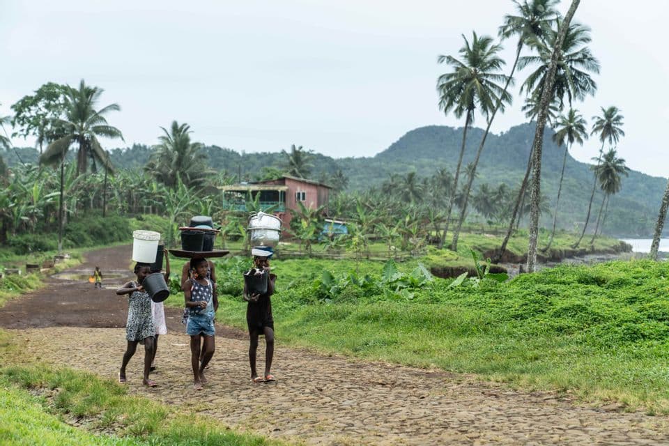 Tre giovani ragazze con secchi e vasi in testa camminano su una strada di ciottoli in un lussureggiante paesaggio tropicale.