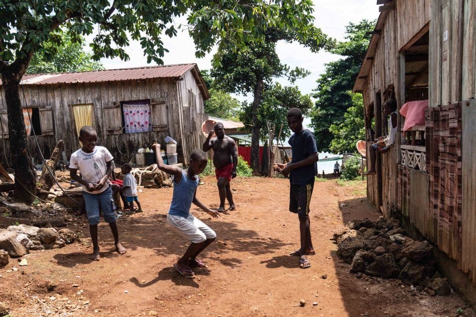 Un gruppo di bambini che giocano e ballano su un sentiero sterrato in un villaggio con semplici case di legno e alberi.