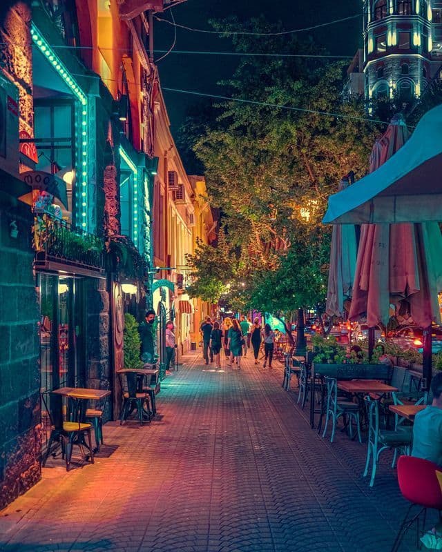 Une rue piétonne étroite, la nuit, bordée de cafés et restaurants en terrasse, illuminée par des néons colorés et des lumières chaleureuses.
