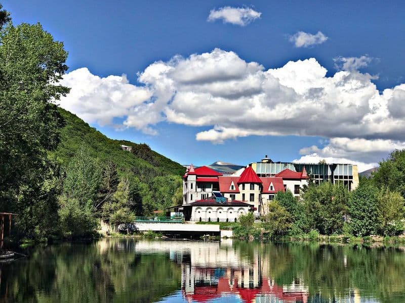Un bâtiment au toit rouge au bord d'un lac se reflète dans l'eau calme, avec une colline boisée derrière sous un ciel nuageux.