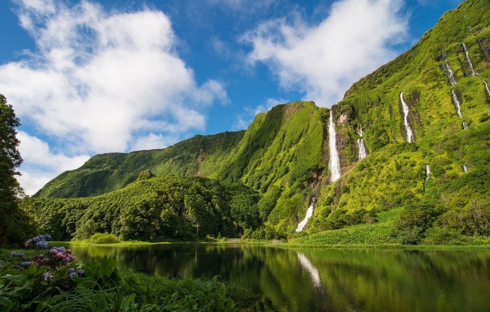 Molteplici cascate scendono da una verde e rigogliosa montagna, riversandosi in un lago calmo che riflette il cielo azzurro e le nuvole.