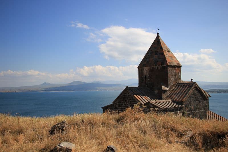 Une église en pierre avec une flèche conique se dresse sur une colline herbeuse sèche dominant un grand lac bleu et des montagnes lointaines.
