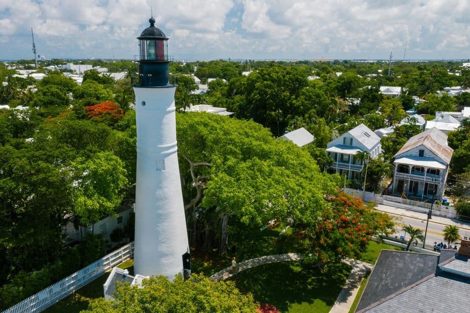 An aerial view of a white lighthouse with a black lantern room towering over a town with lush green trees and houses.