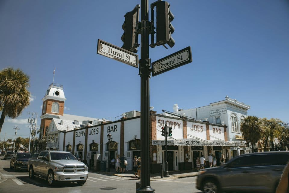 Street signs for Duval St and Greene St on a post at a sunny street corner, with Sloppy Joe's Bar in the background.