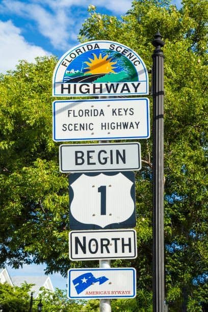 A road sign post indicating the beginning of the Florida Keys Scenic Highway and US Route 1 North, with trees in the background.