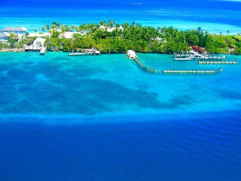 An aerial view of a small tropical island with palm trees and buildings, surrounded by turquoise and deep blue water with wooden docks.
