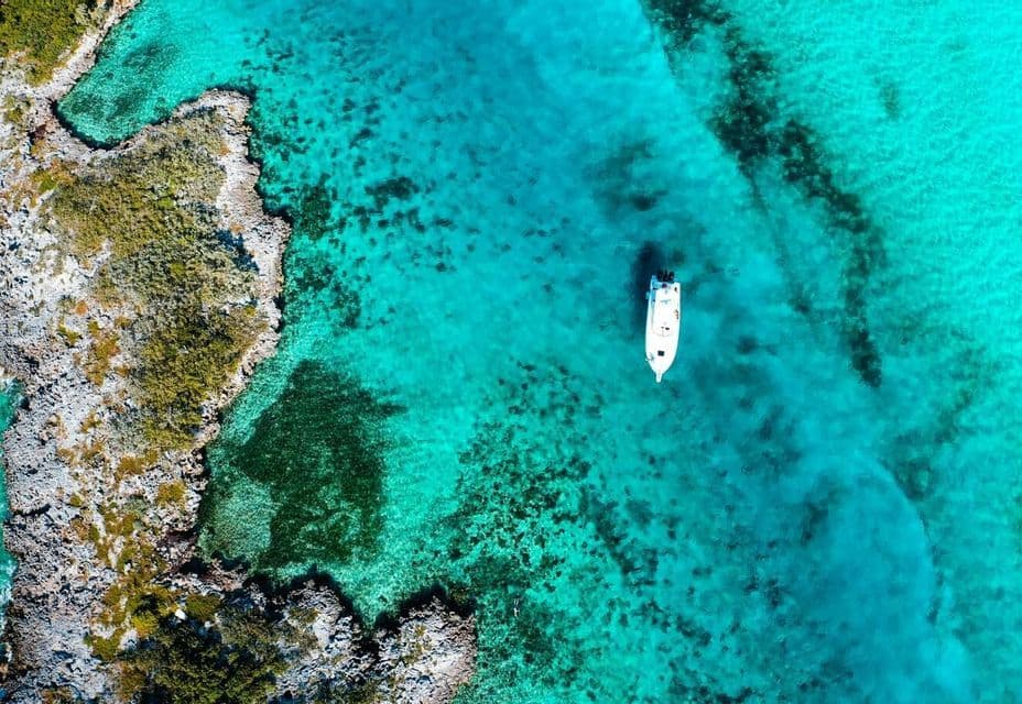 An aerial, top-down view of a white boat in clear, turquoise water beside a rocky coastline with green vegetation.