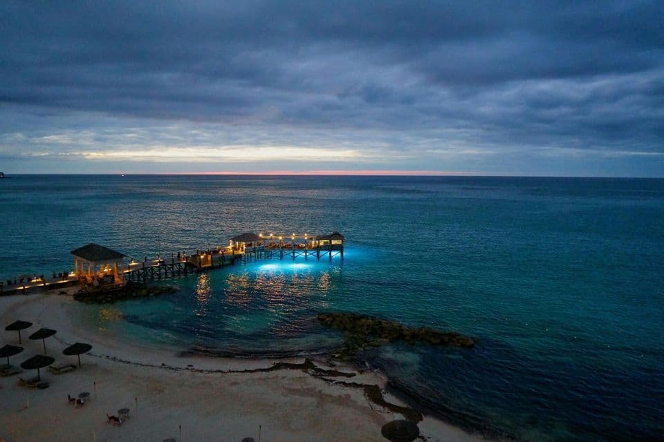 An illuminated wooden pier extends from a sandy beach over calm turquoise water at dusk under a cloudy sky.
