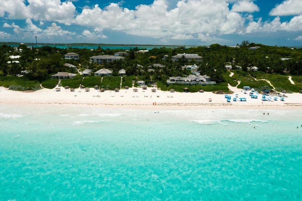 An aerial view of a white sand beach with clear turquoise water, lined with resorts and lush green vegetation under a cloudy blue sky.