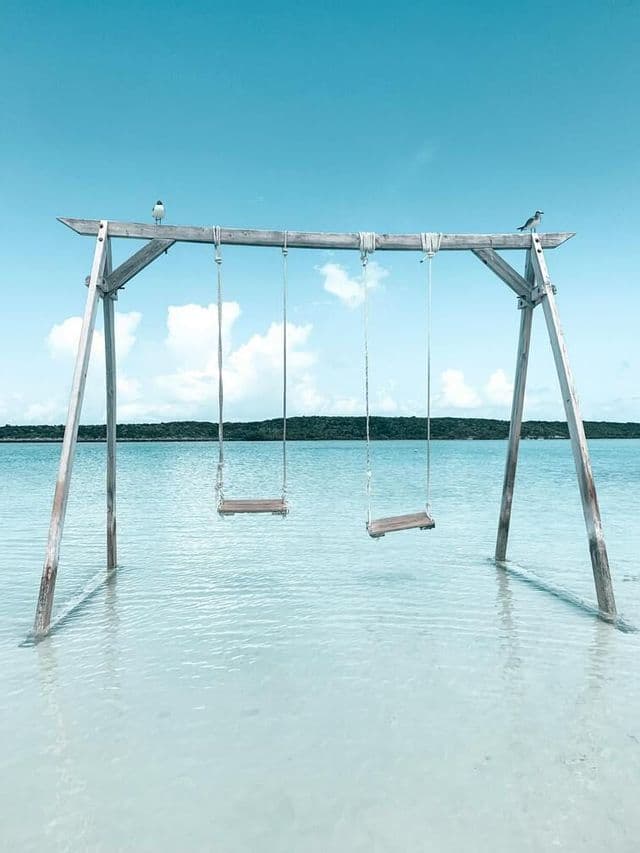 A wooden swing set with two empty swings stands in shallow turquoise water under a blue sky, with two birds perched on top.