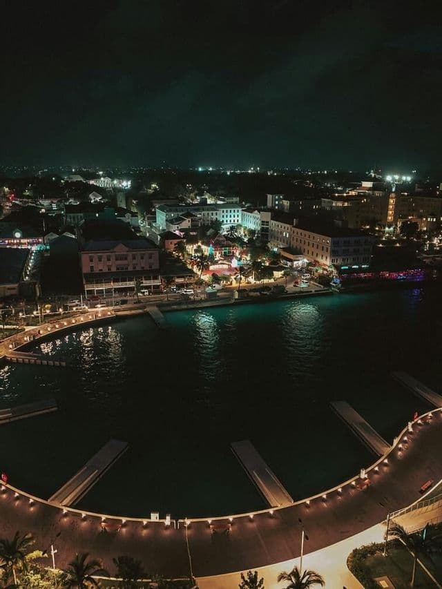 An aerial view of a coastal city and harbor at night, with illuminated buildings and a curved pier in the foreground.