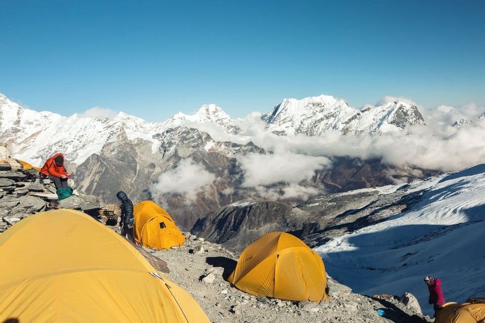 Un groupe WeRoad campe sous des tentes jaunes sur un flanc de montagne rocheux, avec une vaste chaîne de sommets enneigés en arrière-plan sous un ciel dégagé.
