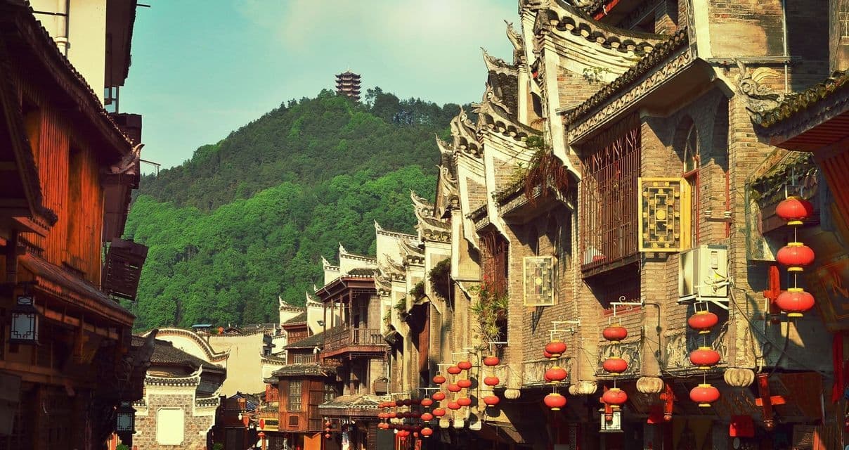 Una calle estrecha con edificios de estilo tradicional y farolillos rojos, con una pagoda visible en una colina boscosa distante.