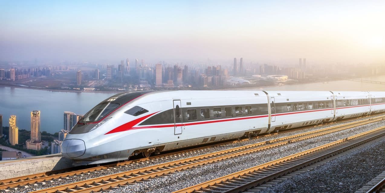 Un tren de alta velocidad en vías férreas con vistas a un horizonte urbano moderno y un río bajo un cielo brumoso.