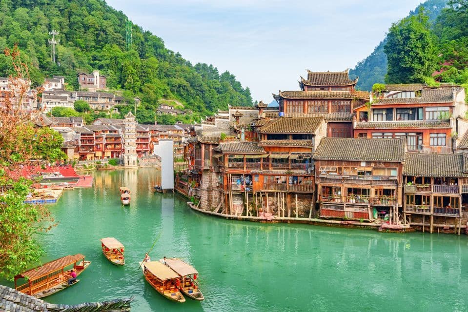 Pequeñas embarcaciones de madera flotan en un río turquesa que atraviesa un pueblo histórico de casas sobre pilotes al pie de una montaña verde.
