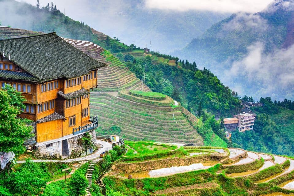 Una casa de madera de varios pisos con vistas a exuberantes terrazas de arroz verdes, construidas en la ladera de una cordillera brumosa.