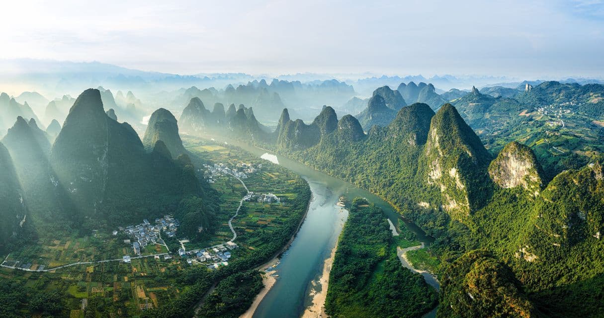 Vista aérea de un amplio río fluyendo por un valle rodeado de verdes montañas kársticas y pequeños pueblos bajo un cielo brumoso.