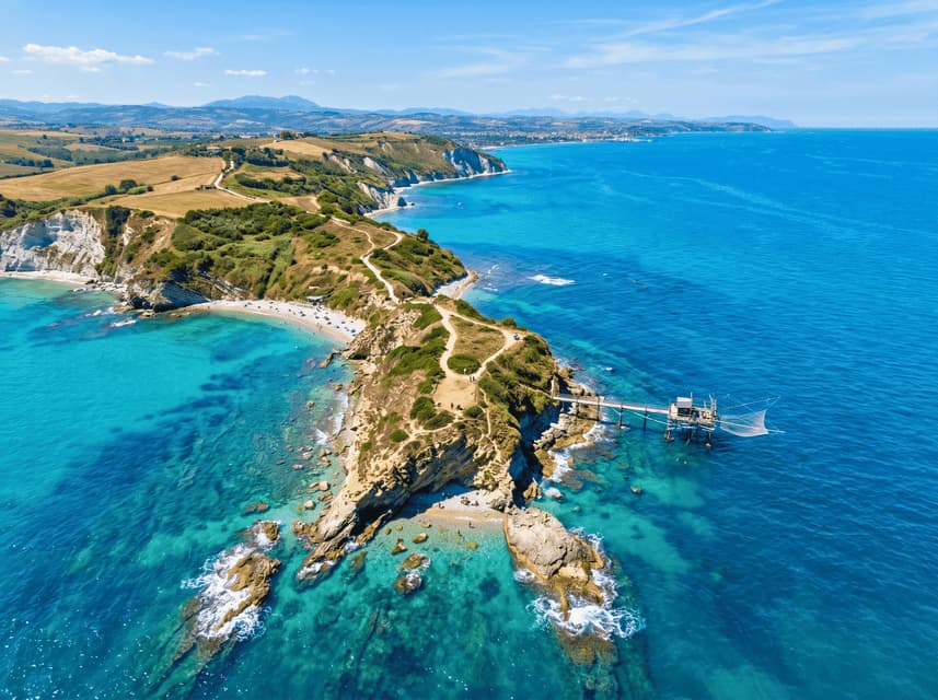 Vista aerea di una costa rocciosa con un tradizionale molo da pesca che si estende nel mare turchese accanto a una piccola spiaggia.