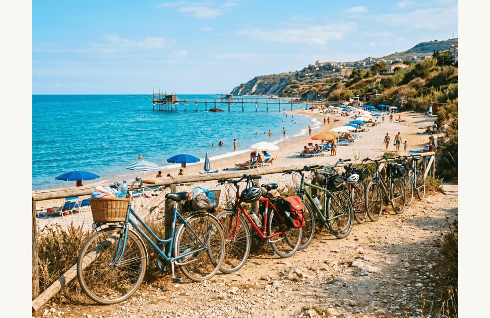 Biciclette allineate lungo un sentiero che si affaccia su una spiaggia soleggiata con un pontile e un mare turchese.