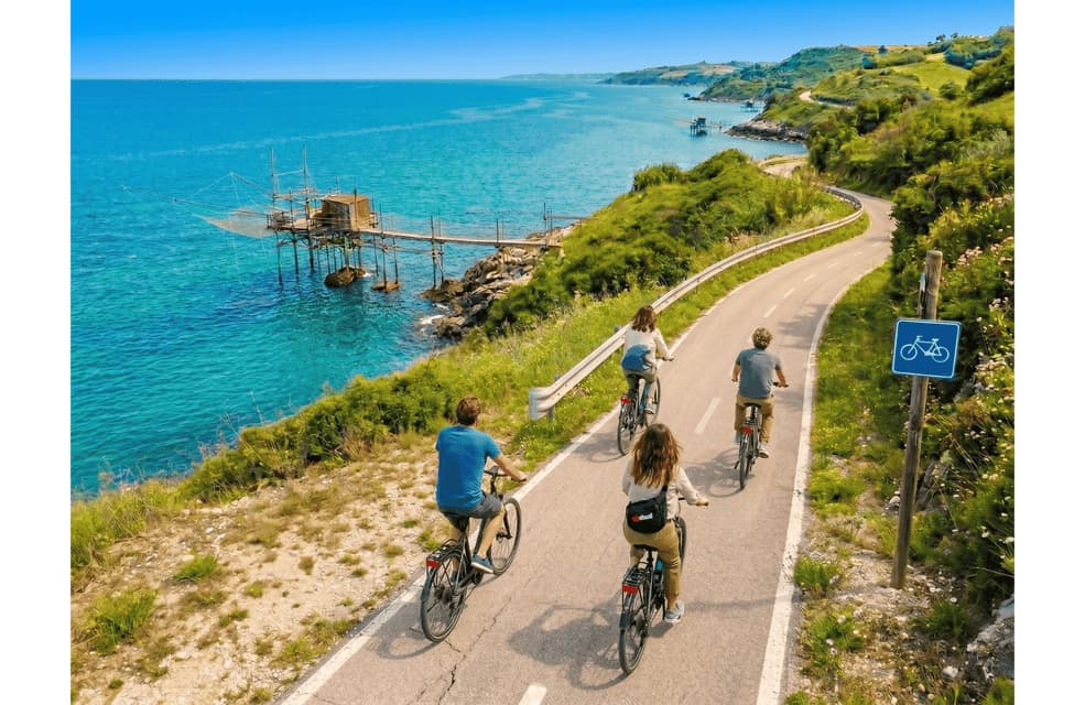 Un viaggio di gruppo WeRoad in bicicletta lungo una strada costiera tortuosa, affacciata su un mare turchese e con un tradizionale molo di pescatori.