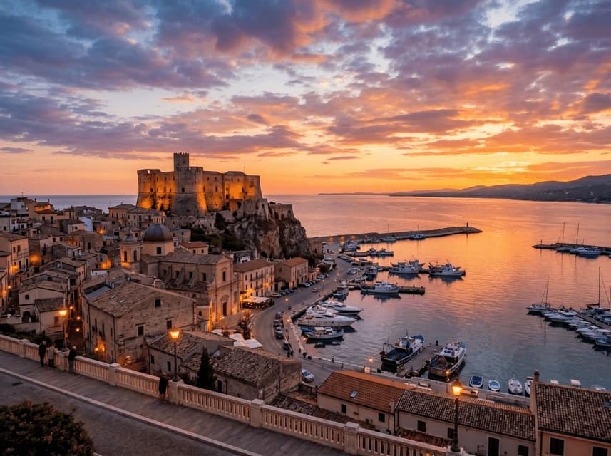 Un castello illuminato su una scogliera si affaccia su una città costiera e un porto pieno di barche al tramonto.