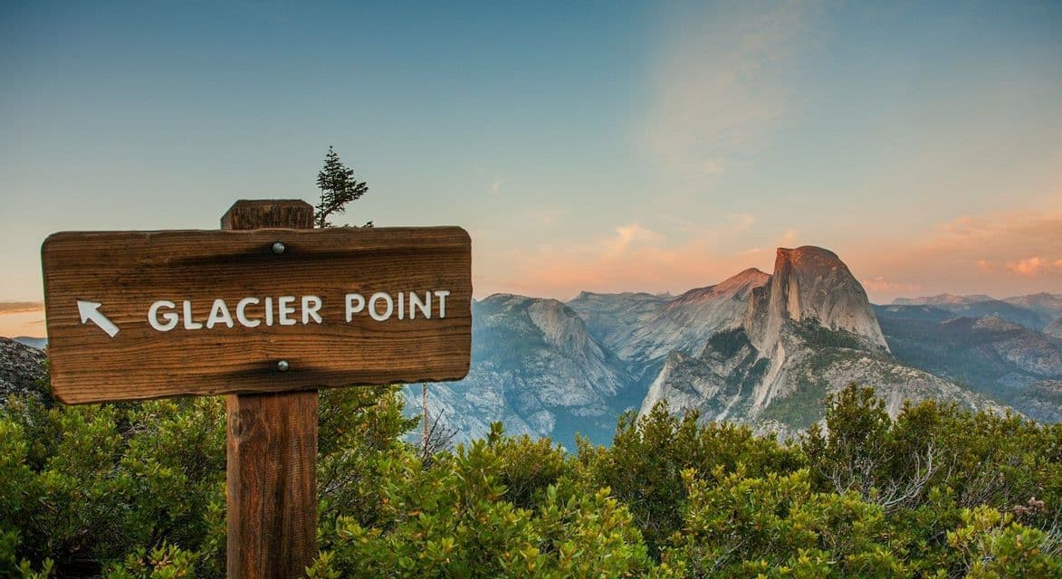 A wooden sign reading 'Glacier Point' overlooks a vast mountain range with a prominent granite dome at sunset.