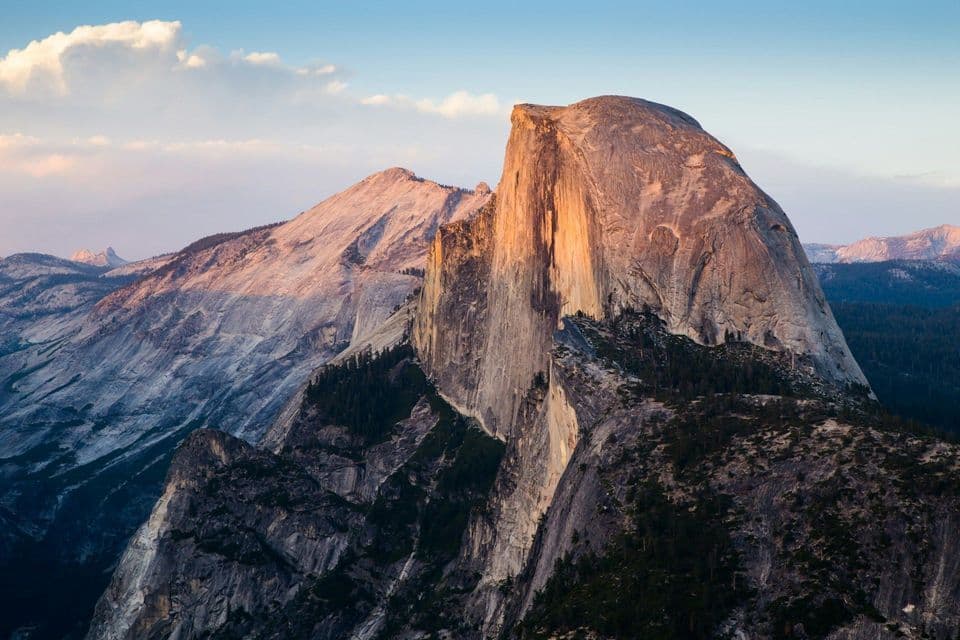 The sheer cliff face of a large granite dome glows with warm sunset light in a vast mountain range under a blue sky with clouds.