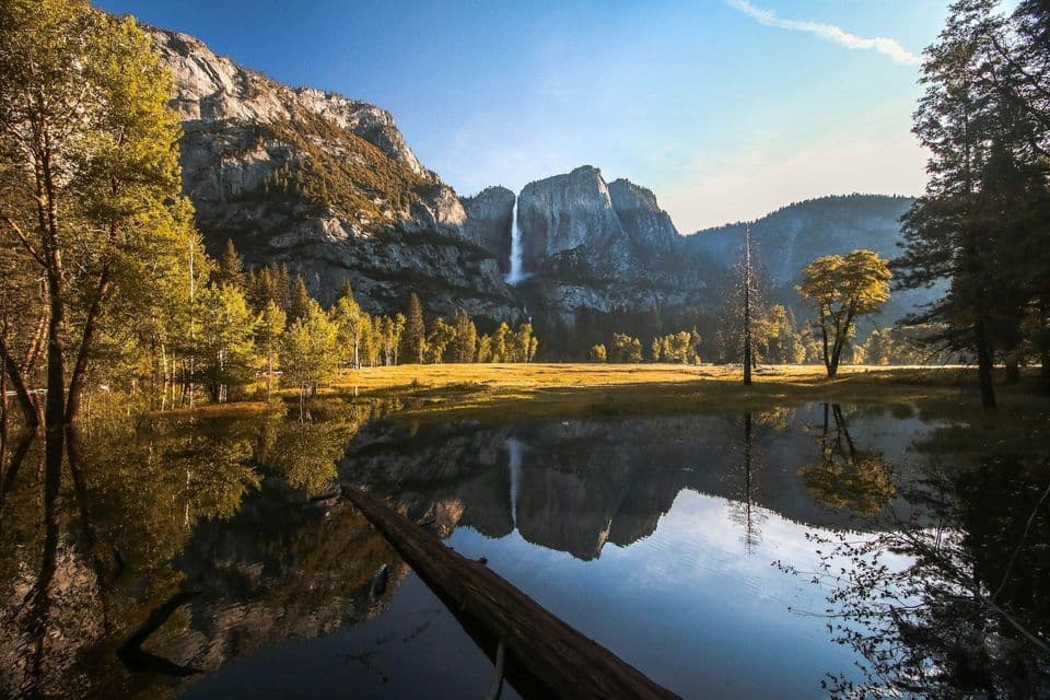 A waterfall on a granite mountain range is reflected in a calm lake, with autumn trees and a meadow in the foreground.