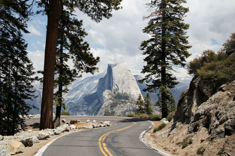 A winding paved road curves through a pine forest, with a view of a large granite dome and mountains under a partly cloudy sky.