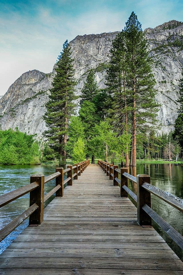 A wooden bridge with railings extends over a river, leading toward a pine forest at the base of a large granite mountain.