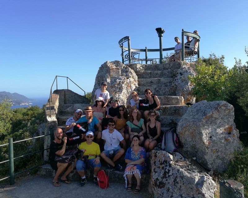 Eine WeRoad-Gruppe posiert für ein Foto auf Steinstufen an einem Aussichtspunkt mit Blick auf das Meer unter einem klaren blauen Himmel.