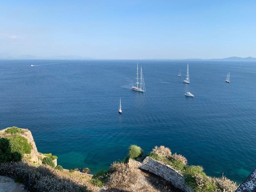 Vogelperspektive von einer felsigen Klippe auf mehrere Segelboote und Yachten auf einem weiten, tiefblauen Meer unter klarem Himmel.