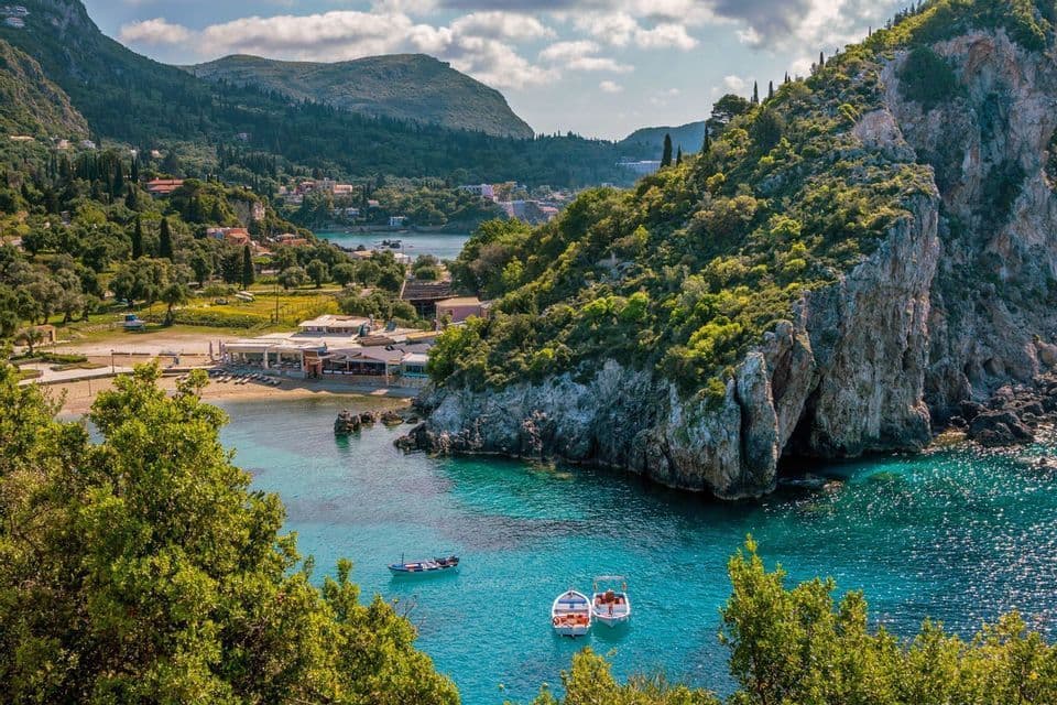 Ein erhöhter Blick auf eine Küstenbucht mit türkisfarbenem Wasser, kleinen Booten, einem Sandstrand und üppig grünen Klippen.