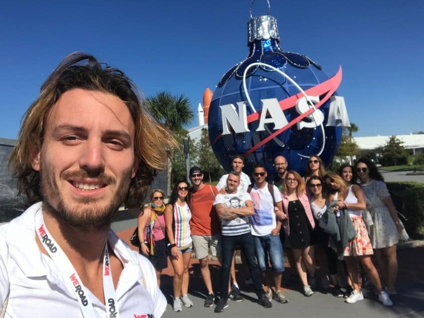 Un grupo de viaje de WeRoad se toma una selfie frente a un gran adorno con el logo azul de la NASA bajo un cielo despejado.