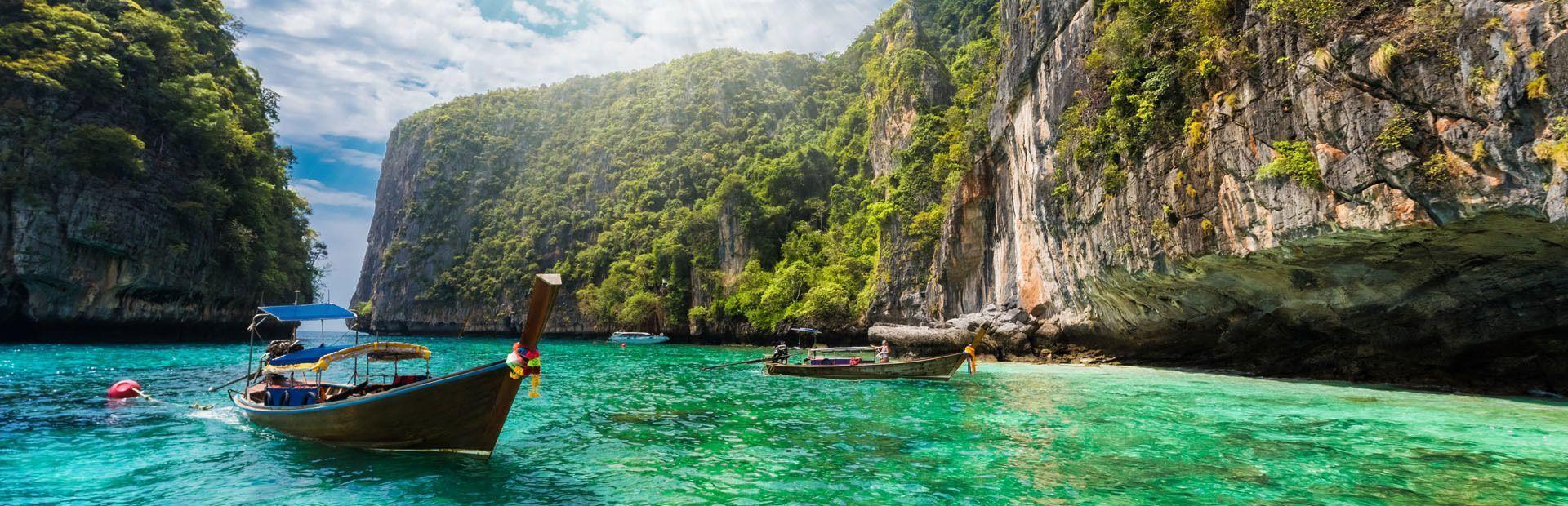 Traditional long-tail boats float in a turquoise lagoon surrounded by tall, green-covered cliffs under a partly cloudy sky.