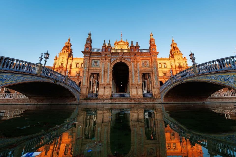 An ornate brick building with two tiled bridges is reflected in the water below during a clear sunset.