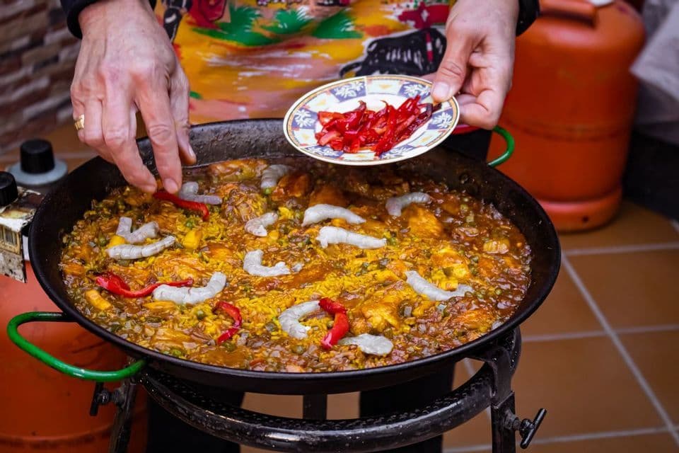A person's hands adding sliced red peppers and shrimp to a large pan of paella cooking outdoors on a burner.