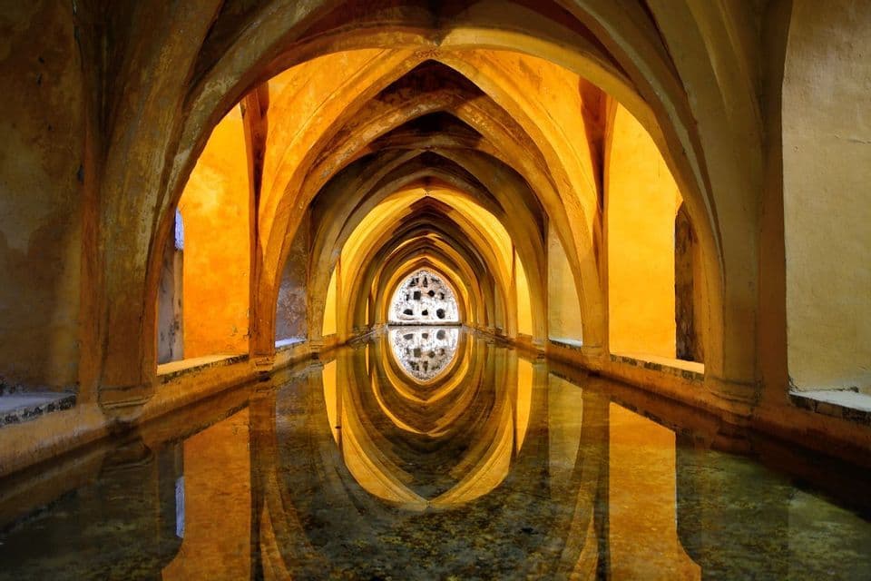 Golden arches lining a long hall are reflected in a central pool of water, creating a symmetrical, tunnel-like view.