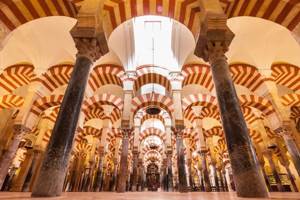 The interior of a large hall filled with rows of dark columns supporting a ceiling of red-and-white striped arches.