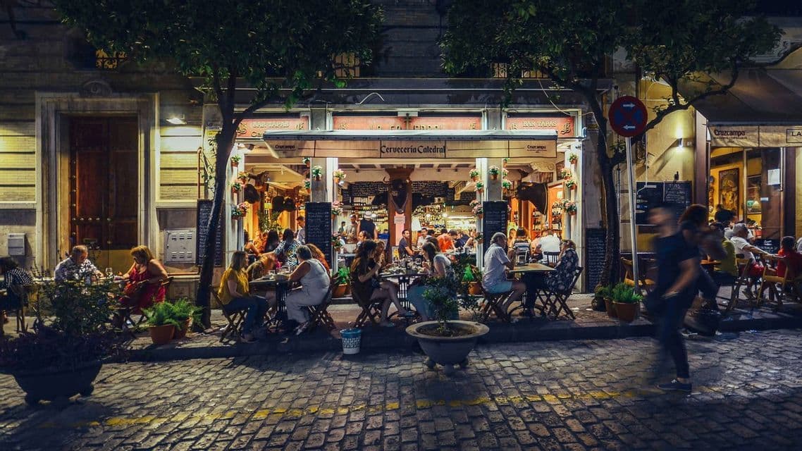 People dining at the outdoor tables of a busy restaurant on a cobblestone street at night.
