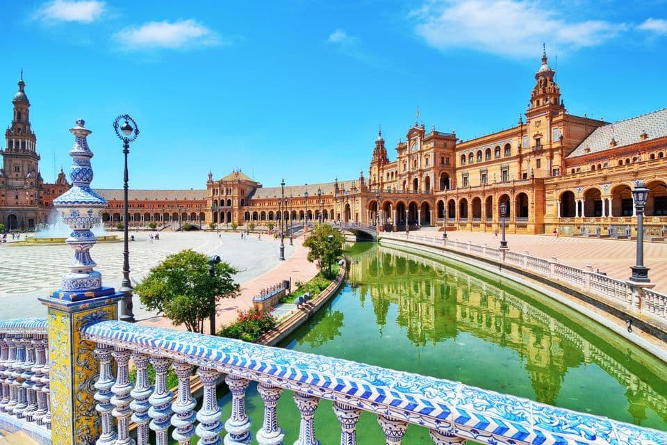 An ornate building with towers and arches is reflected in a canal, viewed from a bridge with a blue and white tiled railing.