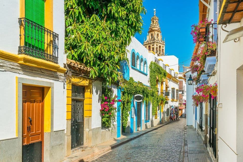 A narrow cobblestone street is lined with colorful whitewashed buildings, with a historic bell tower visible in the distance under a clear blue sky.