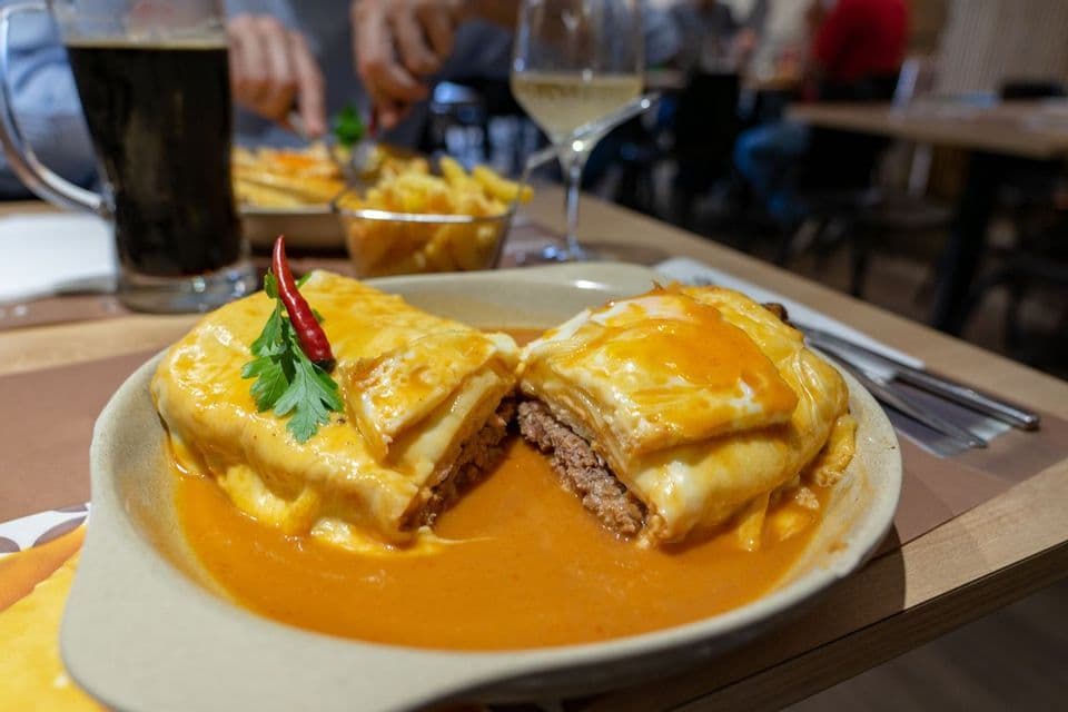 A meat-filled sandwich cut in half, topped with melted cheese and a fried egg, served in a bowl of orange sauce on a table.