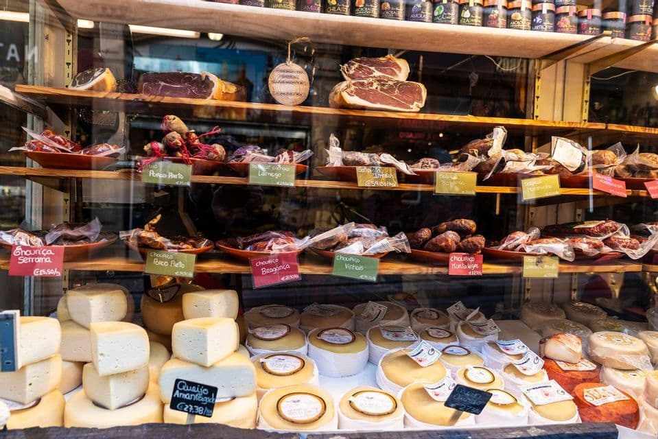 A variety of cheeses and cured meats with price tags displayed on wooden shelves in a delicatessen window.