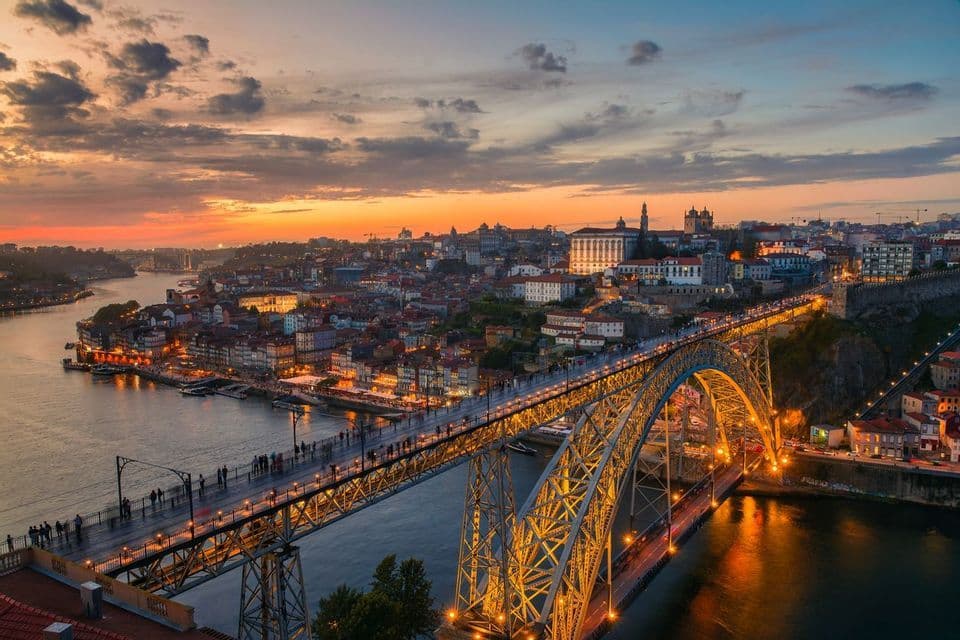 An illuminated double-deck arch bridge with people walking on it spans a river in a cityscape at sunset.